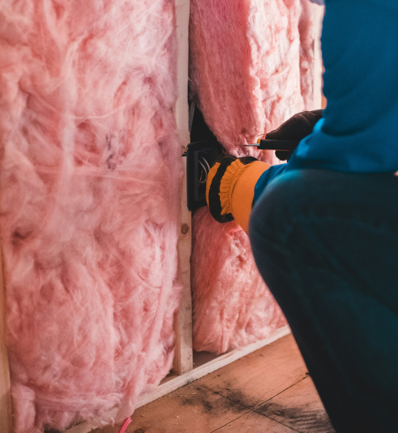 Person is blue overalls with orange gloves sitting on wooden fllor with insulation. Credit: Erik Mclean